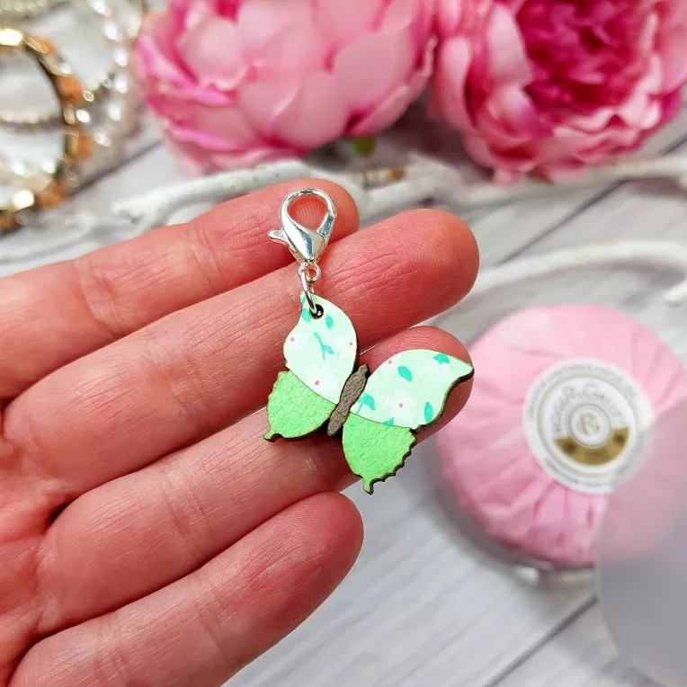 Green wooden stitch marker on hand against a background with pink flowers