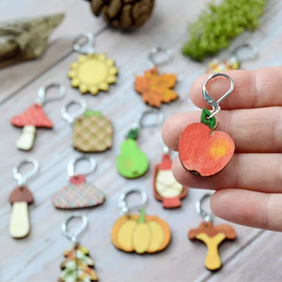 Apple, a handpainted wooden stitch marker, held on hand with autumnal stitch markers in background